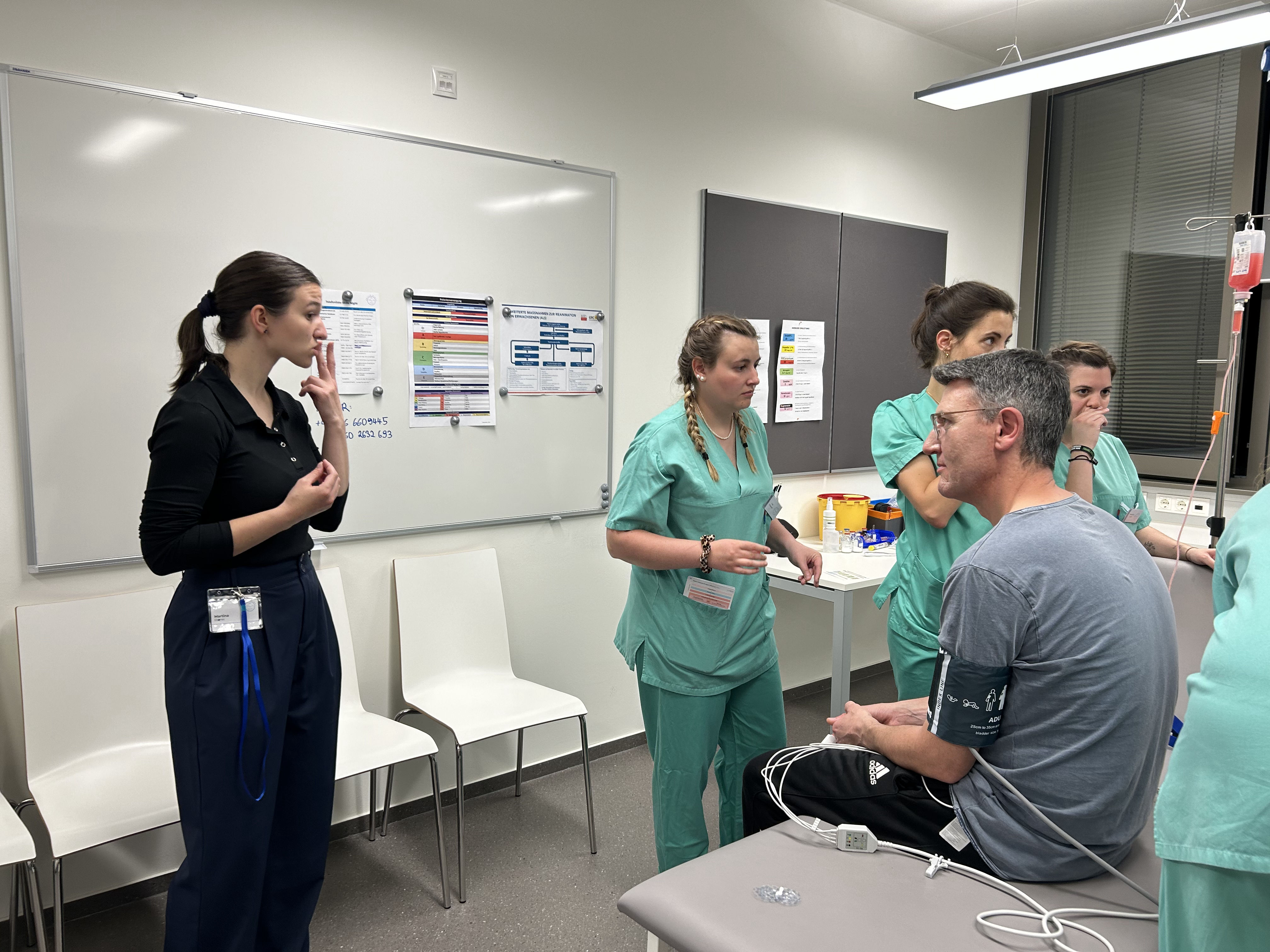 Eine Studierende spricht in Gebärdensprache mit einem Darsteller, der auf einer Liege sitzt, Um ihn sind Studierende mit der medizinischen Versorgung beschäftigt.