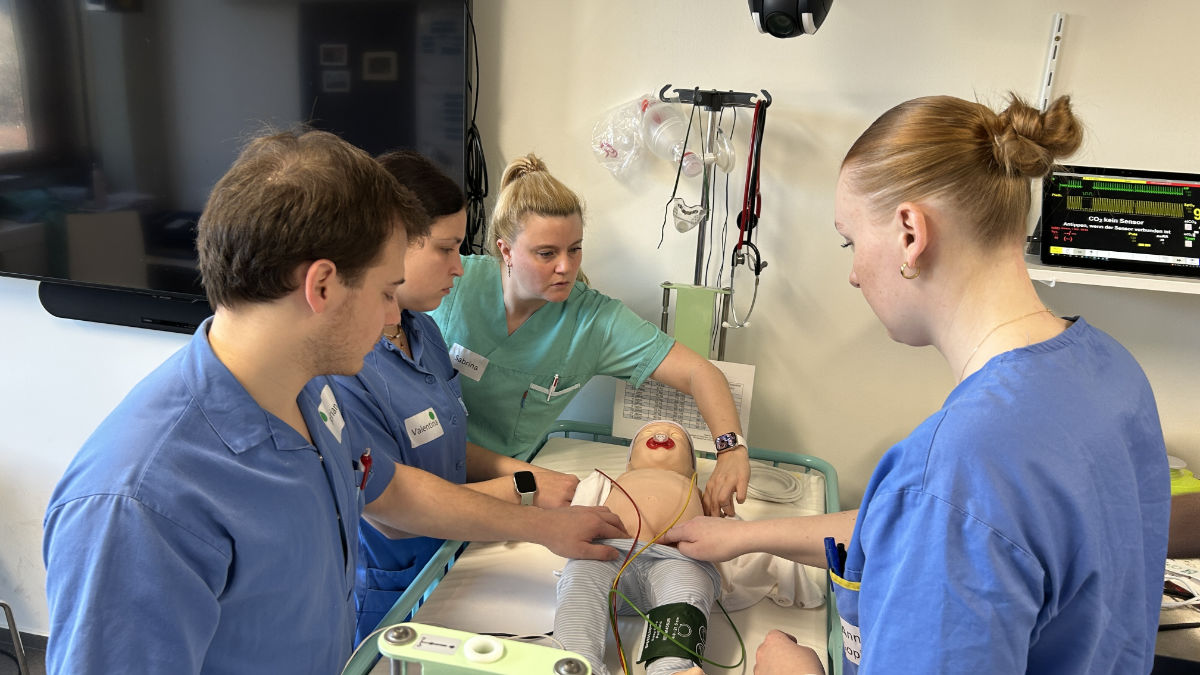 Studierende messen einer Säuglings-Simulationspuppe den Puls, eine Lehrende unterstützt sie