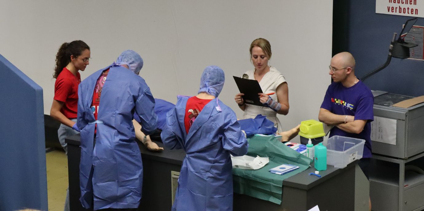 Zwei Personen in blauer OP-Kleidung arbeiten in einem Hörsaal an einer Puppe, beobachtet von zwei Aufsichtspersonen